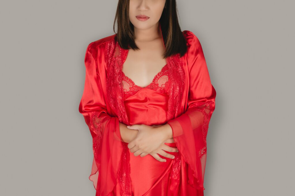 A woman in a nightgown and a satin red robe wakes up in the middle of the night because of menstrual pain. Female sleepless because of stomach aches on a gray background.